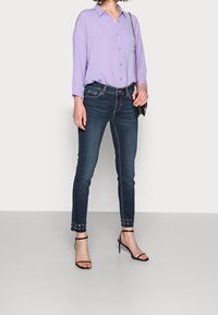 Ljuslila skjorta med knappar och ficka, tillsammans med mörkblå smala jeans med nitar vid fållen och svarta remsandaler med klack.