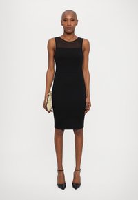 Black sleeveless bodycon dress with a sheer neckline, smooth texture, knee-length cut, paired with a gold clutch and black high heels.