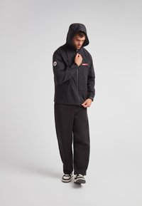 Giacca nera impermeabile con cappuccio, chiusura con zip, tasca anteriore e un distintivo logo rosso e bianco sulla manica, abbinata a pantaloni neri larghi.