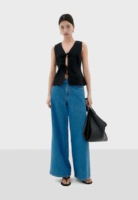 Frau in einem ärmellosen schwarzen Oberteil mit vorderen Bindebändern, weiten blauen Jeans, schwarzen Sandalen und hält eine große schwarze Handtasche vor einem schlichten Hintergrund.