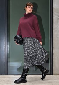 Burgunderfarbener Rollkragenpullover, bedruckter schwarz-weißer Midirock und knielange schwarze Lederstiefel; hält eine schwarze Handtasche.