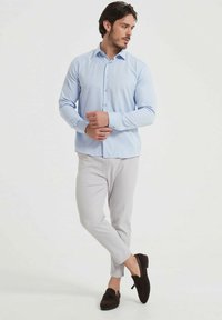 Chemise bleu clair à manches longues avec un devant boutonné, associée à un pantalon texturé gris clair et des mocassins marron foncé, en pose debout.