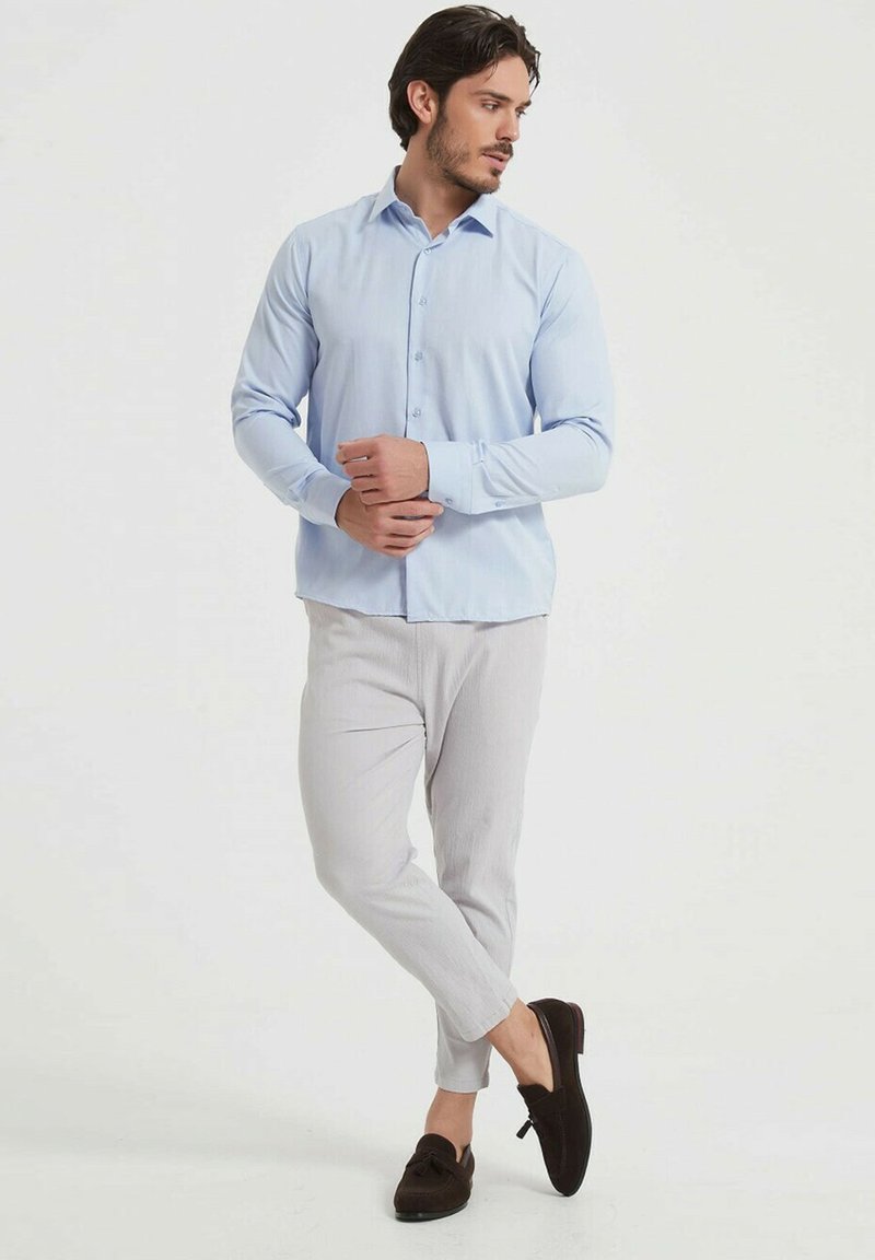 Chemise bleu clair à manches longues avec un devant boutonné, associée à un pantalon texturé gris clair et des mocassins marron foncé, en pose debout.