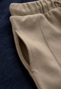 Pantalon de survêtement beige en tissu doux, avec une taille élastique, des poches latérales et une texture lisse, posé sur un fond bleu foncé.