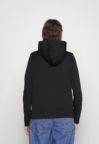 Sudadera con capucha negra, amplia en la parte trasera y con una capucha grande. Cuenta con mangas largas y una pretina acanalada. Combinada con vaqueros azules.