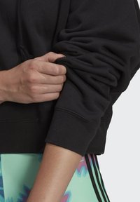 Svart sweatshirt med en mjuk struktur, gathers på ärmarna och ribbade manschetter; en hand som greppar ärmen illustrerar passform och komfort.