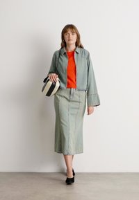 Kvinne som har på seg et sett med grønn denimjakke og skjørt med en oransje topp, holder en svart og hvit veske, går på et grått gulv.