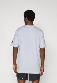 T-shirt en coton gris clair avec une coupe décontractée, des manches courtes et un col rond, présentant un petit logo bleu marine sur la manche gauche.