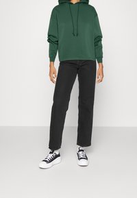 Grön huvtröja med dragkedja, i kombination med svarta raka jeans och svartvita sneakers. Slät tygtextur.