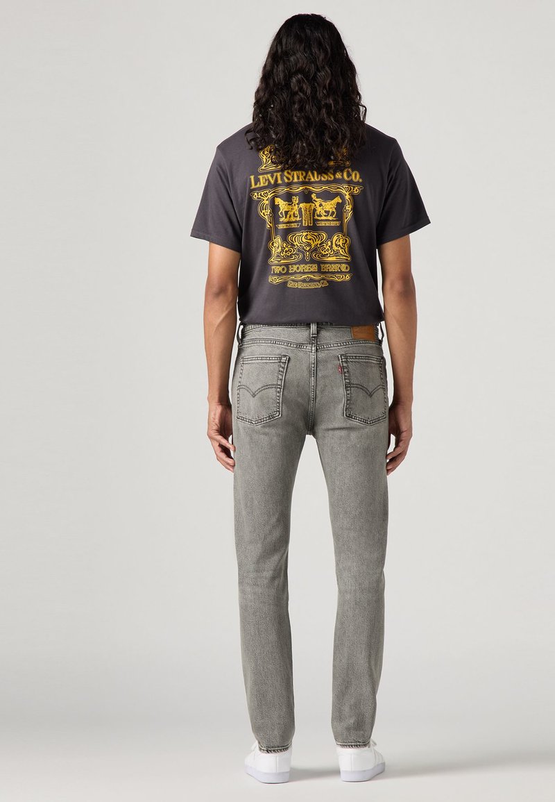 Jean en denim gris avec une coupe droite, présentant une étiquette en cuir marron, et un t-shirt noir avec un imprimé graphique jaune au dos.