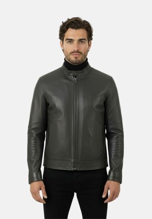 Homme portant une veste en cuir noire avec une fermeture éclair à l'avant et un col montant, associé à un pantalon noir, debout devant un fond blanc.