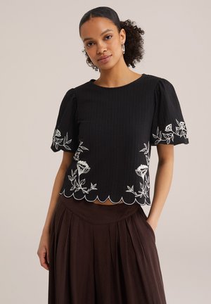 Žena kovrčave kose nosi crnu majicu kratkih rukava s bijelim cvjetnim vezom i valovitim rubom u kombinaciji s smeđom plisiranom suknjom.