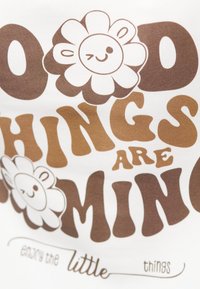 Hvit stoff med brun typografi og smilende blomstergrafikk. Teksten sier "Gode ting kommer" og "nyt de små tingene."