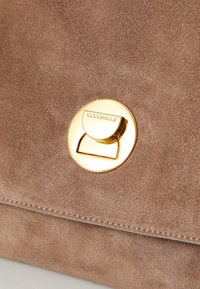 Brown suede handbag with a smooth texture. Features a round gold hardware clasp with "Coccinelle" embossed. Subtle stitching detail along edges.