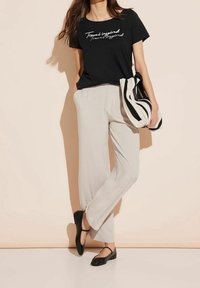 Černé tričko s krátkým rukávem a bílým nápisem, spárované s světle šedými kalhotami s širokými nohavicemi a černými plochými botami, držící černobílou pruhovanou kabelku.