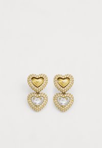 Boucles d'oreilles en forme de cœur en or avec une pierre claire au centre, entourée de strass transparents. Texte "GUESS" visible sur le haut du cœur.
