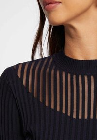 Svart ribbad tröja med genomskinlig randig panel, med en rund halsringning. Tyget har en slät yta med växlande ogenomskinliga och transparenta sektioner.