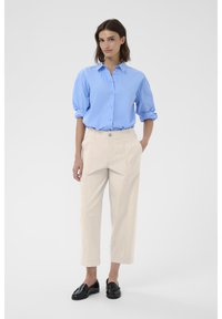 Chemise bleu clair à manches retroussées, pantalon cropped couleur crème avec poches avant, et mocassins noirs au fini brillant.