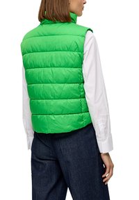 Helle grüne, ärmellose Steppweste mit horizontalen Nähten. Sie hat einen hohen Kragen und wird über einem weißen Langarmshirt und dunklen Hosen getragen.