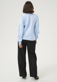 Blouse bleu clair avec un motif en zigzag ; manches longues et bouffantes ; associée à un pantalon noir à jambes larges et des chaussures à talons noires ; fond uni.