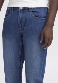 Sötétkék farmernadrág egyenes szabással, finom kopással, öt zsebbel és kontrasztos sárga varrással a varratok mentén.