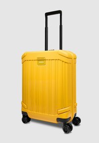 Valise rigide jaune vif à coque dure avec quatre roues pivotantes noires et poignée télescopique noire étendue sur fond gris uni.