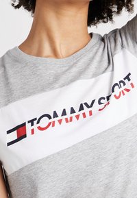 Šedé bavlněné tričko s bílým vodorovným pruhem s nápisem „TOMMY SPORT“ v tučném červeném a černém provedení. Kulatý výstřih, krátké rukávy.