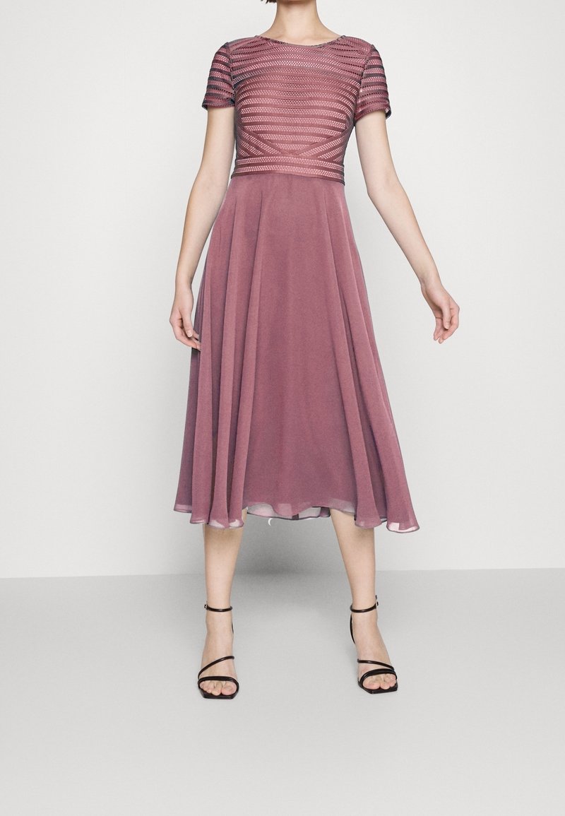 Ein mauve Kleid mit einem strukturierten, taillierten Oberteil mit horizontalen Streifen und einem fließenden, knielangen Rock. Kombiniert mit schwarzen Riemchenschuhen.