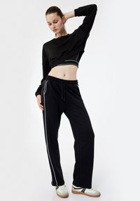 Kobieta w czarnej krótkej bluzie, czarnych spodniach z białymi paskami i białych sneakersach, pozująca z jedną ręką za głową na białym tle.