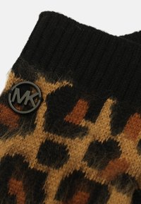 Pull en tricot avec un col noir côtelé, un motif géométrique brun et beige, et une étiquette ronde noire avec "MK" sur le côté.
