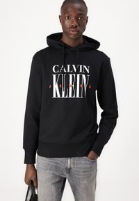 Juodas medvilnės džemperis su kapuce, ant kurio yra baltos ir oranžinės spalvos užrašas "CALVIN KLEIN JEANS". Su raišteliais apyrankės ir juosmens, derinamas su šviesiai pilkais džinsais.
