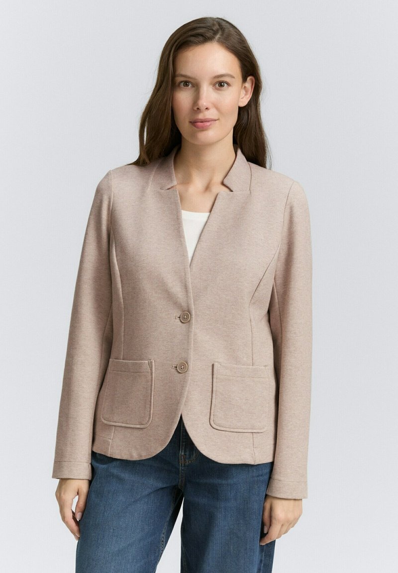 Blazer beige chiaro in un morbido tessuto texturizzato. Presenta un colletto a punta, due tasche frontali e una chiusura con due bottoni. Abbinato a jeans blu.