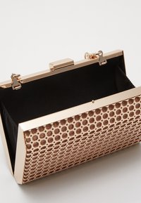 Metaliczna złota kopertówka z wzorem plastra miodu, o strukturalnym kształcie, zamknięciem na zatrzask oraz czarną podszewką wewnątrz. Krawędzie wykończone na wysoki połysk.