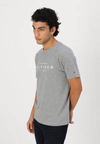 Grå bomull t-skjorte med korte ermer, rund hals, med hvit trykt "TOMMY HILFIGER EST. 1985" logo og en liten logo-patch på ermet.