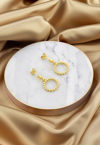 Guldörhängen med en pärldesign, visade på en rund vit marmorplatta med en gyllene kant, mot en bakgrund av satin.