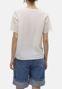 T-shirt blanche avec des manches courtes brodées en œillets, associée à un short en denim bleu à ourlet retroussé. Vue arrière simple, texture de tissu douce.