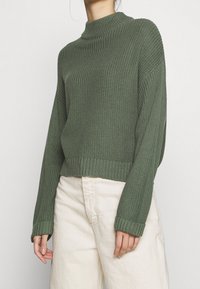 Zielony dzianinowy sweter z wysokim kołnierzem, prążkowaną teksturą i długimi rękawami, charakteryzujący się krótkim krojem i jednolitym kolorem.