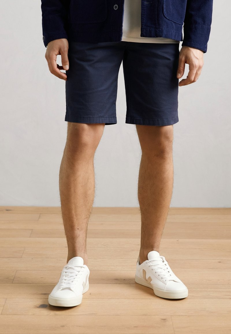 Homme portant un short bleu marine, des baskets blanches et une veste bleu foncé, debout sur un sol en bois devant un mur uni.