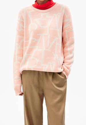 Personne portant un pull en maille rose clair avec des motifs blancs abstraits, un col roulé rouge en dessous et un pantalon beige ample avec une main dans la poche.