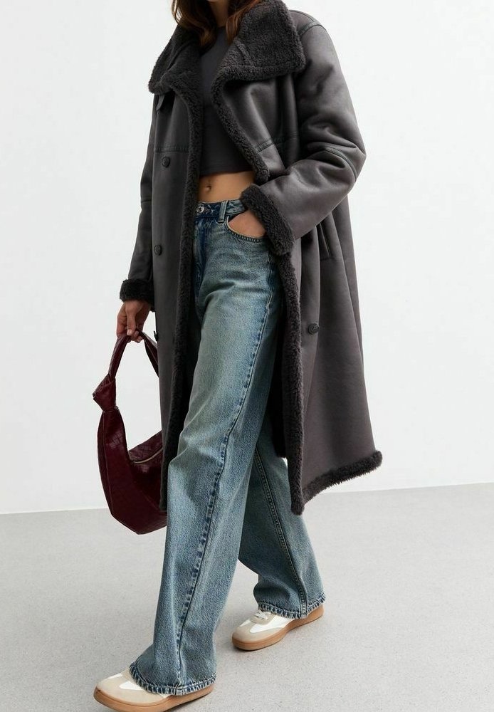 Lungo cappotto grigio con colletto e rifiniture in pelliccia sintetica, indossato sopra un top corto grigio e abbinato a jeans blu chiaro e scarpe con plateau color crema.