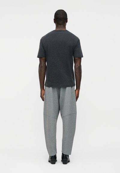 Άνδρας που στέκεται με την πλάτη γυρισμένη, φορώντας μαύρο κοντομάνικο μπλουζάκι, άνετο γκρι παντελόνι και μαύρα παπούτσια, μπροστά σε απλό λευκό φόντο.