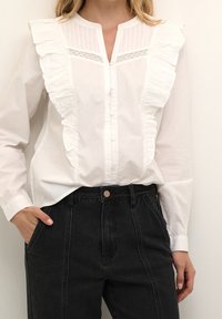 White button-up shirt with ruffled detail along the left side and pinstriped texture at the collar. Black high-waisted jeans.