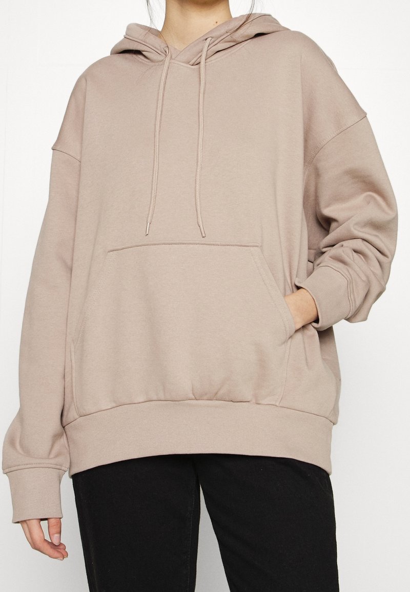 Personne portant un sweat à capuche beige oversize avec poche avant et cordons, associé à un pantalon noir, une main dans la poche sur un fond uni.
