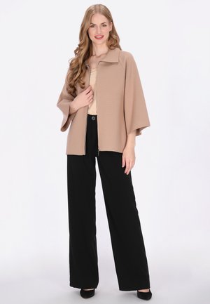 Béžová bunda na zip s límcem, volným střihem a širokými rukávy, doplněná černými kalhotami s širokými nohavicemi. Hladká textura, minimální designové detaily.