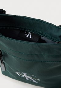 Zöld nejlon táska cipzáras nyílással, fekete belsővel és látható címkével. Fehér logóval és varrási részletekkel díszítve.
