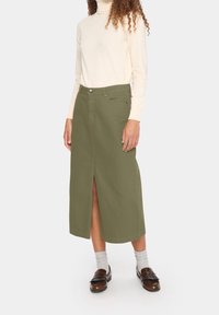 Falda larga verde oliva con una abertura frontal, hecha de un tejido resistente. Combinada con un suéter de cuello alto de manga larga beige y mocasines marrones.