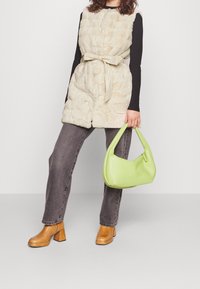 Beżowa kamizelka futrzana z paskiem do wiązania, czarna bluzka z długim rękawem, szare dżinsy, beżowe botki na słupku oraz jasna, zielona torebka o strukturze.