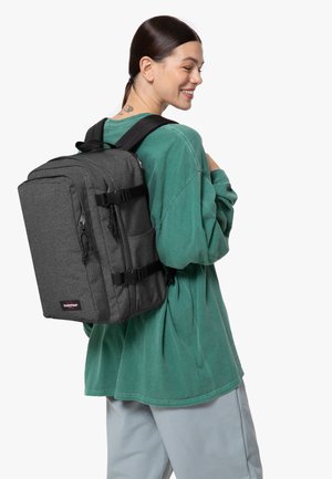 Zaino rettangolare grigio con spallacci regolabili, cerniere e fibbie frontali, indossato sopra una felpa oversize verde e pantaloni chiari.