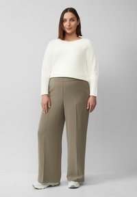 Witte, langarm trui gecombineerd met wijde, olijfgroene broek. De outfit wordt aangevuld met lichtgekleurde sneakers.