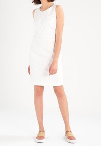 Robe blanche sans manches à silhouette ajustée, col rond et texture lisse, accompagnée de chaussures slip-on tan et blanches.